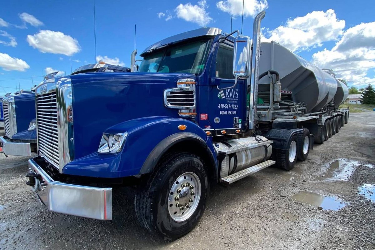 Glossy blue Freightliner with a logo on its door beside a silver tanker trailer and a cloudy sky.