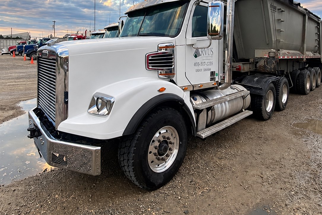 White tractor with an aluminum dump trailer under a cloudy dusk sky.