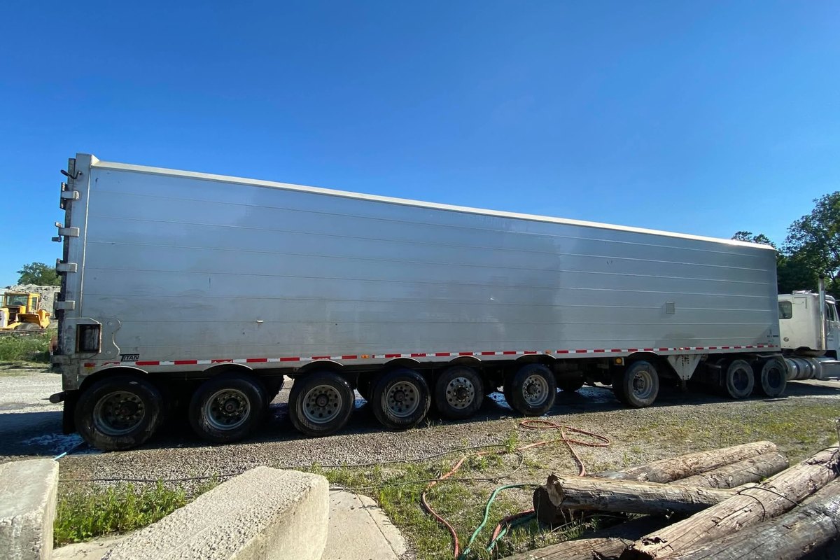 Long silver aluminum trailer with bright even panels on gravel, with logs and concrete blocks in the foreground.