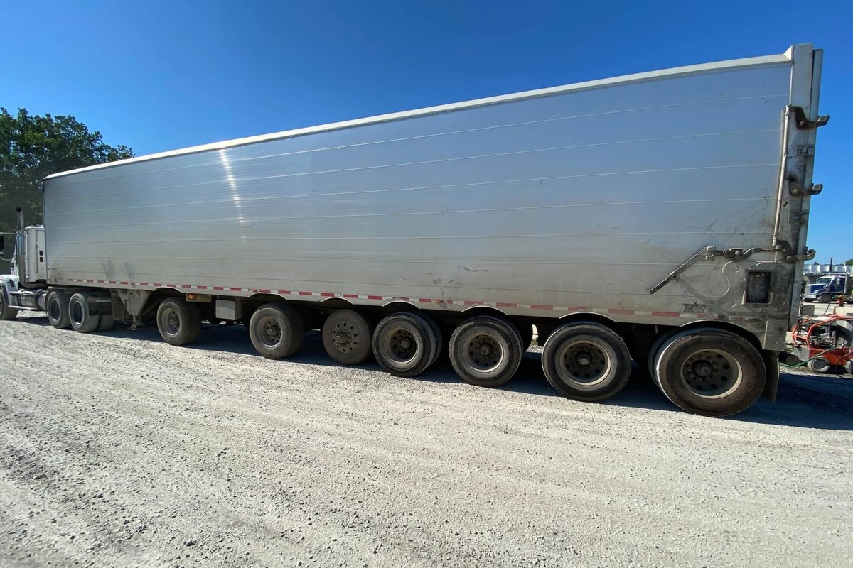 Long silver aluminum trailer on gravel under a bright blue sky with seven wheels along the bottom.