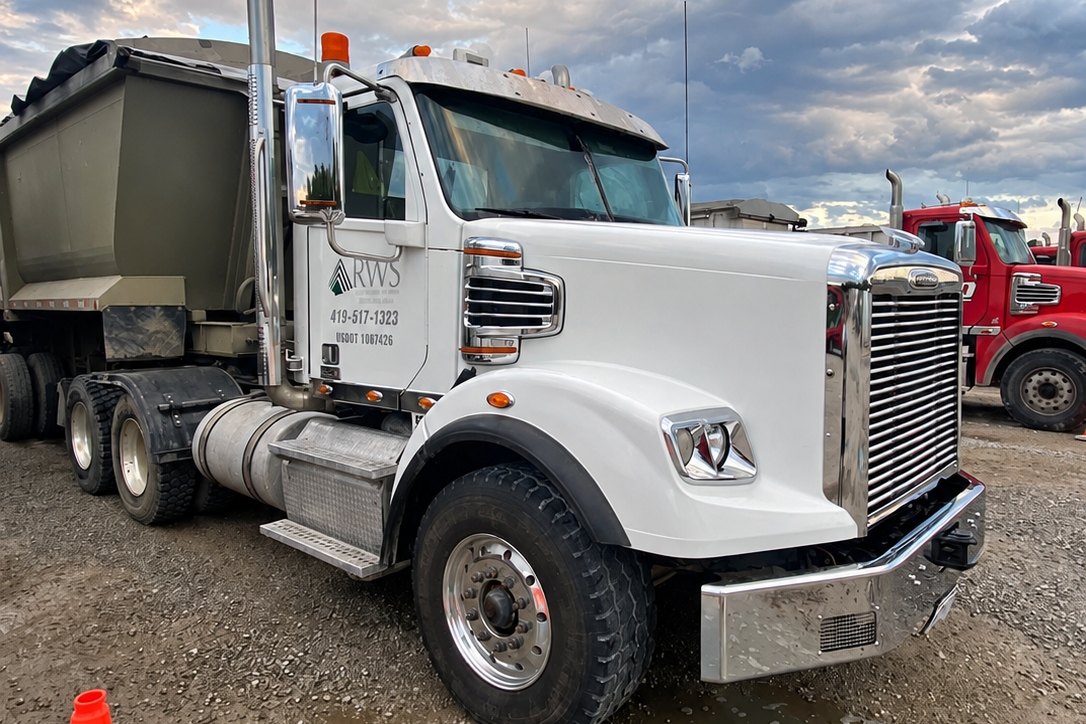 White Freightliner tractor and gray dump trailer from a front-quarter angle, with a chrome grille, bumper and silver tank.