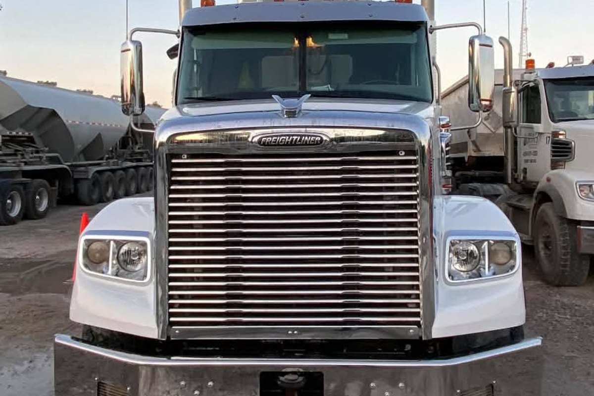 Head-on view of a clean white Freightliner tractor with a bright chrome grille, bumper and mirrors on wet gravel.