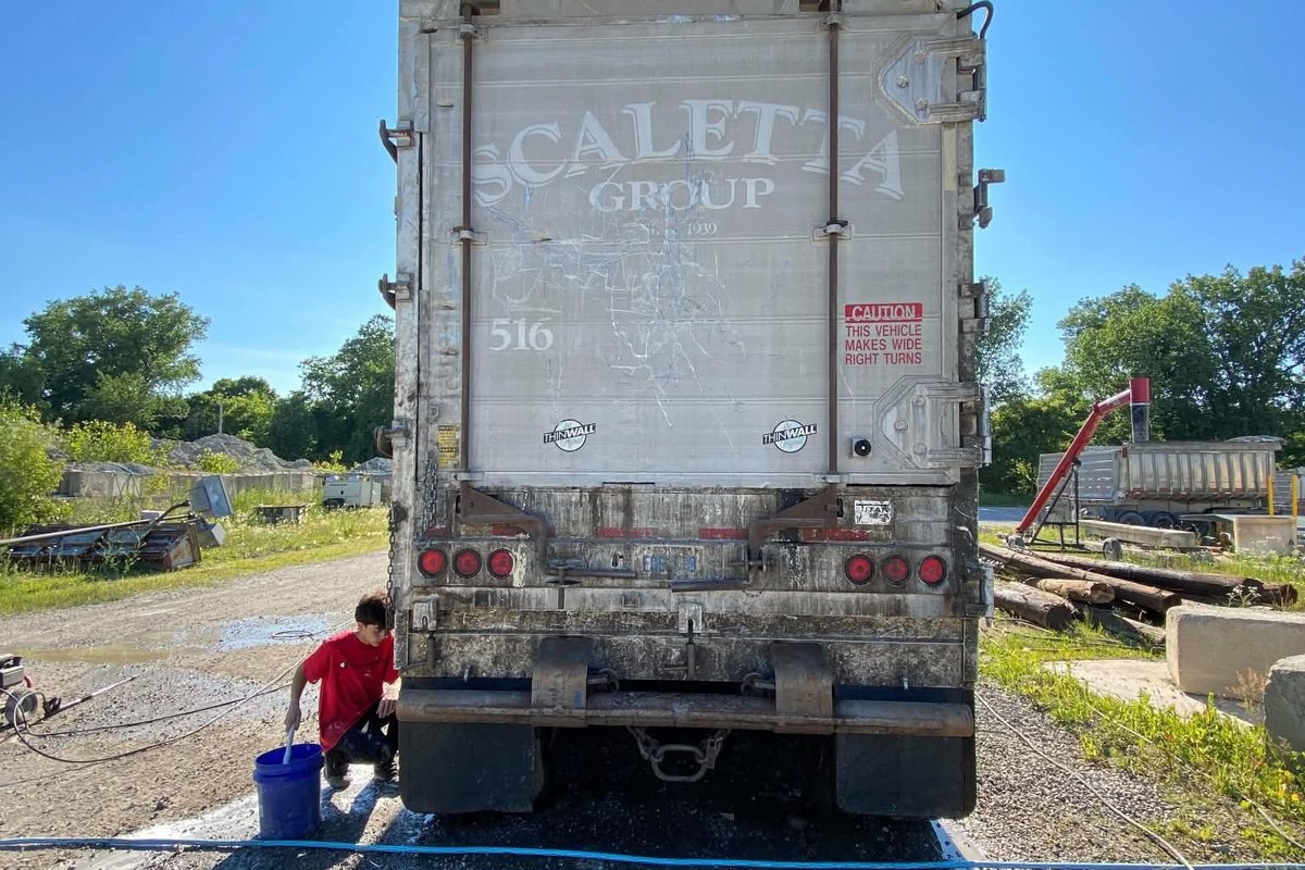 Back of a dirty aluminum trailer with one person kneeling at the left to scrub it, a bucket and a hose beside them.