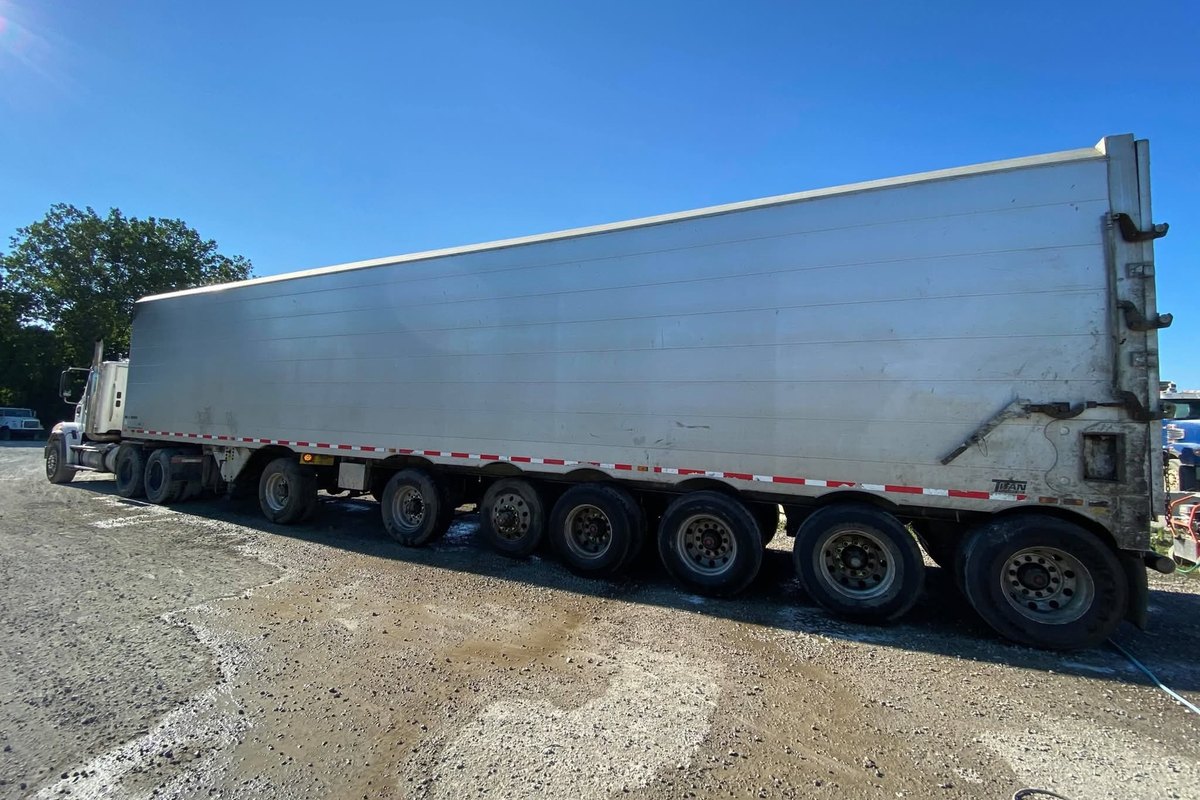 Long aluminum trailer with a white tractor at its front on gravel with trees behind it, seen from the side.