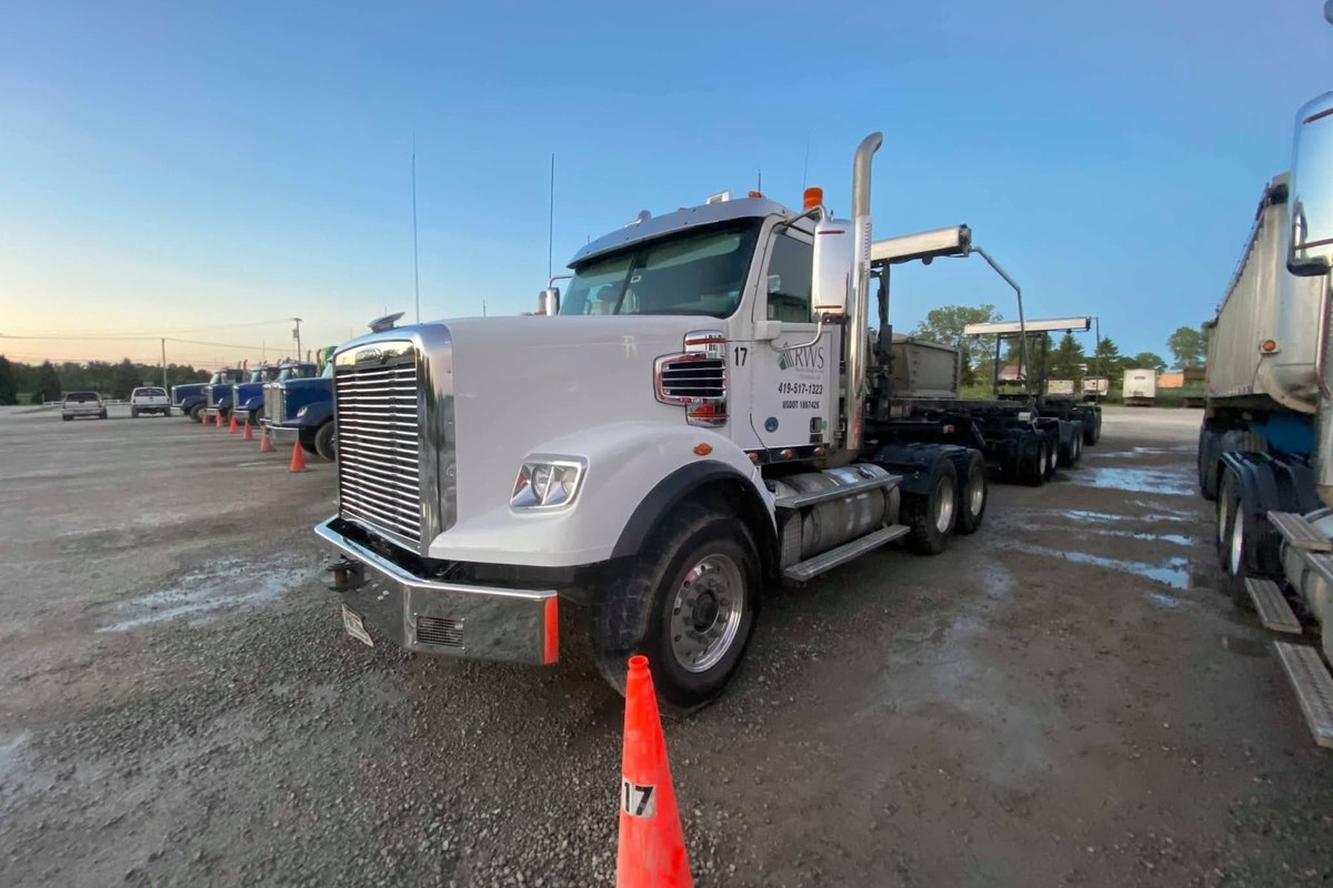 Clean white tractor with a polished wheel, chrome stack and bumper and a roll-off trailer behind it at dusk.