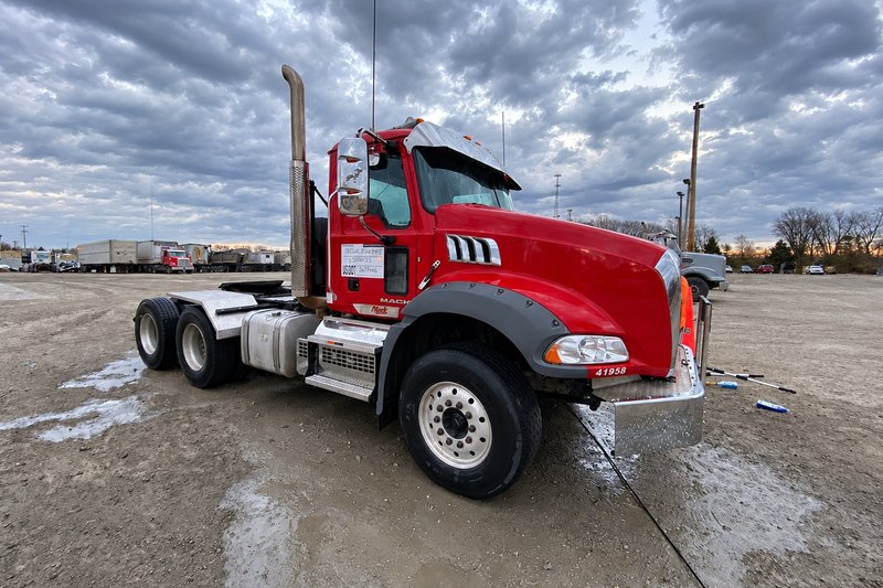 Clean red Mack tractor with silver wheel rims, steps and fuel tank on wet gravel, with parked trailers behind.