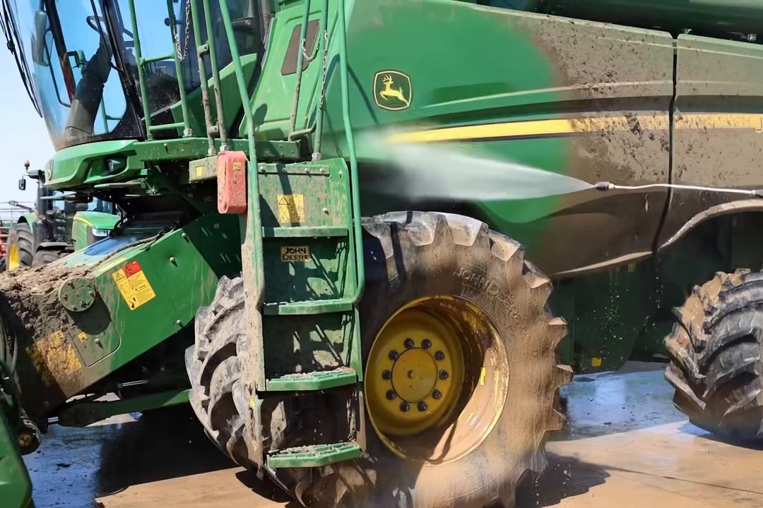 Green combine with yellow wheels being pressure washed, part of the side panel rinsed clean and mud on the tires.