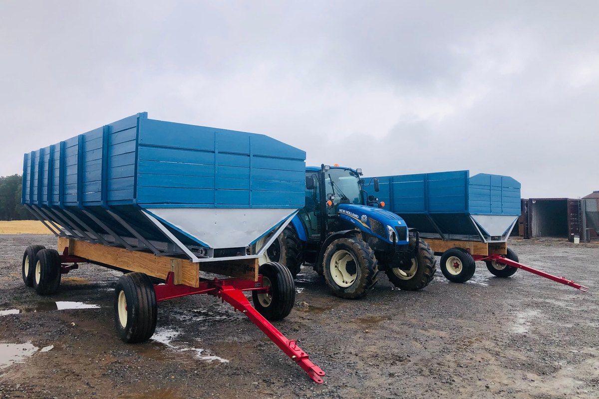 Two blue grain wagons with silver hoppers parked on a wet gravel farm yard beside a blue tractor.