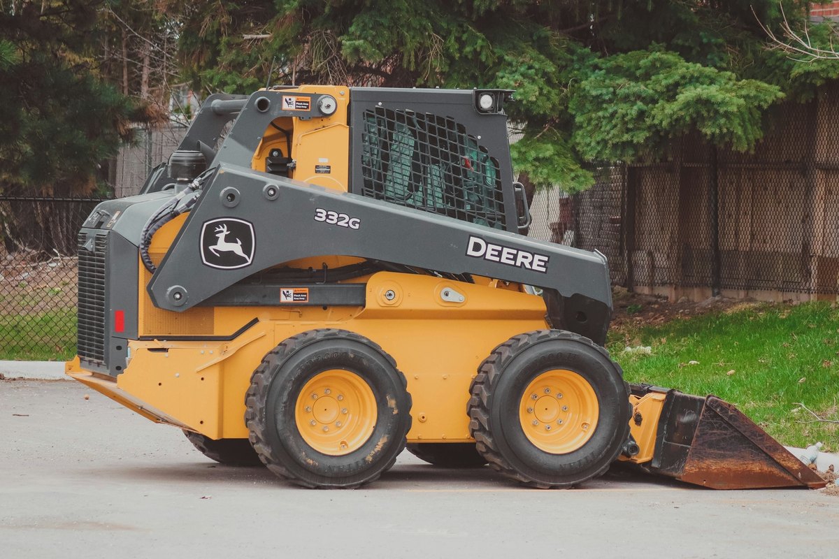 Yellow and gray skid steer loader parked on a paved lot with its bucket down, evergreen trees and a fence behind.
