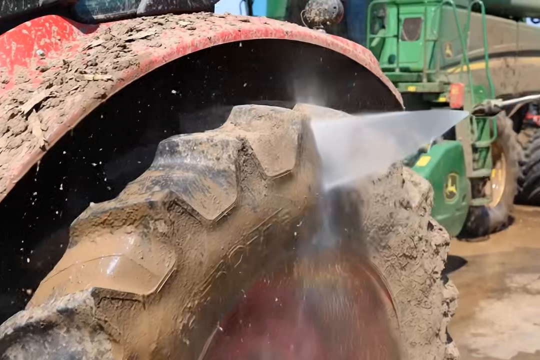 Muddy tractor tire under a red fender being hit by pressure washer spray, with a green combine parked behind.