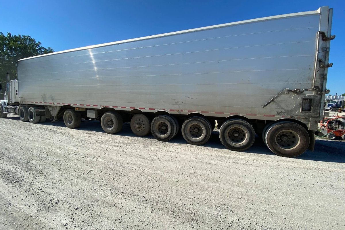 Side of a long silver aluminum trailer standing on gravel under a bright blue sky, with trees behind.