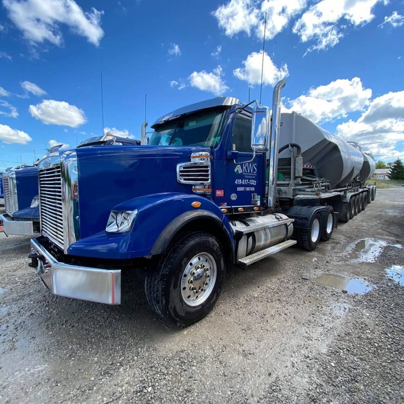 Blue tractor with a company logo on the door and a silver tanker trailer behind it, under a sky of white clouds.